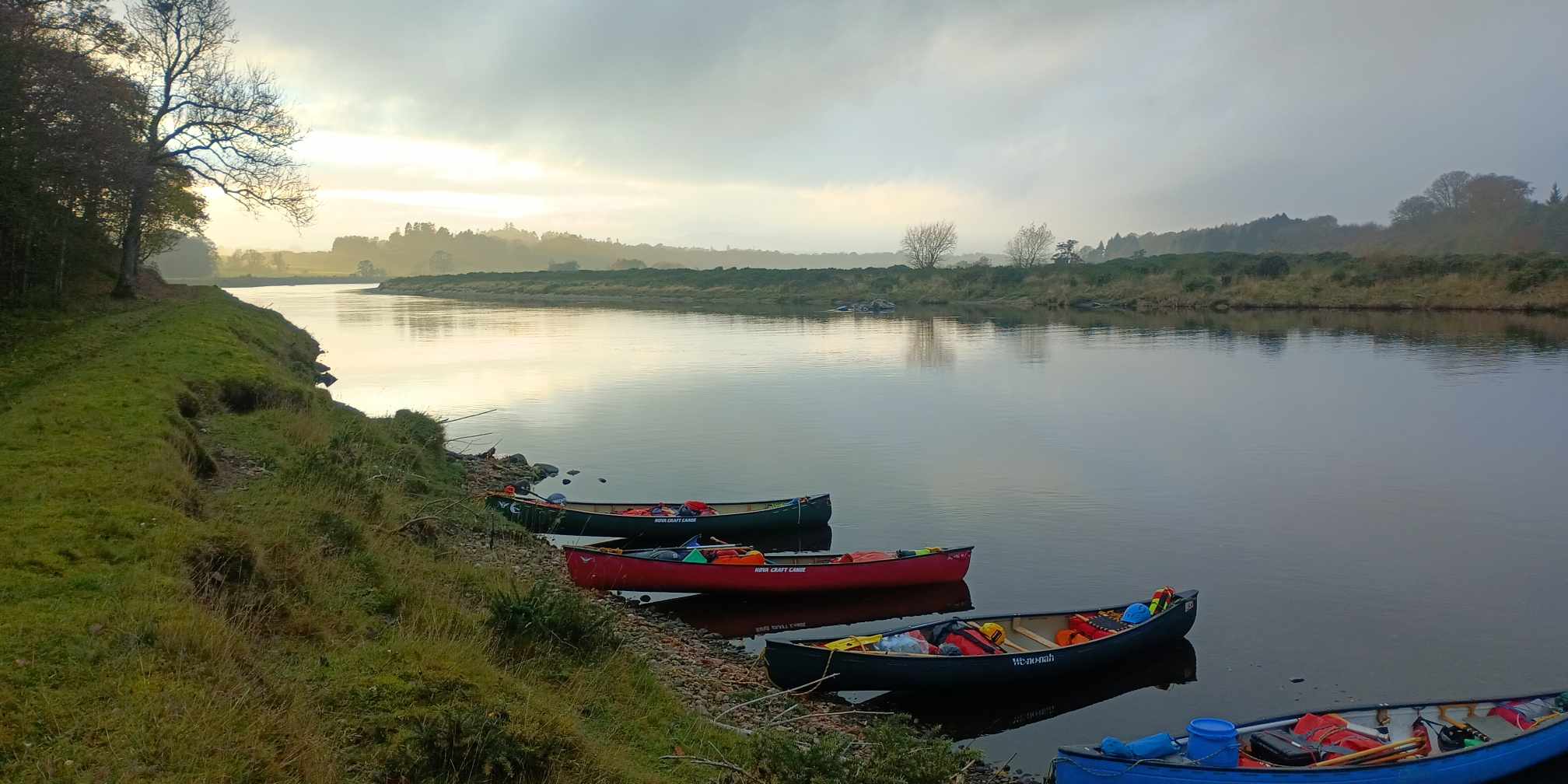 Line of canoes on misty river at dawn with wooded banks