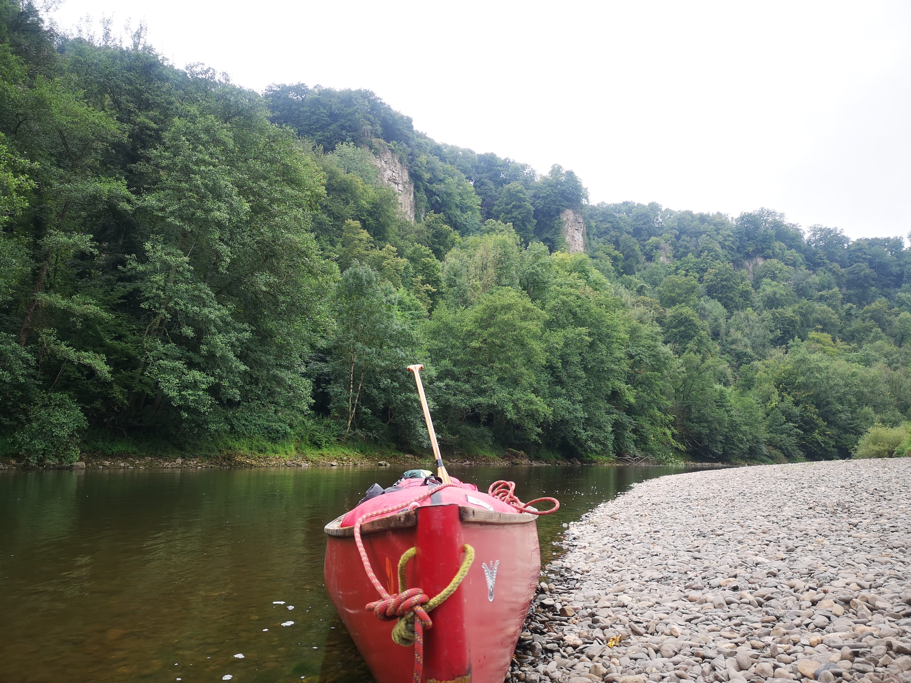 Red canoe resting on riverbank with forested cliffs in background
